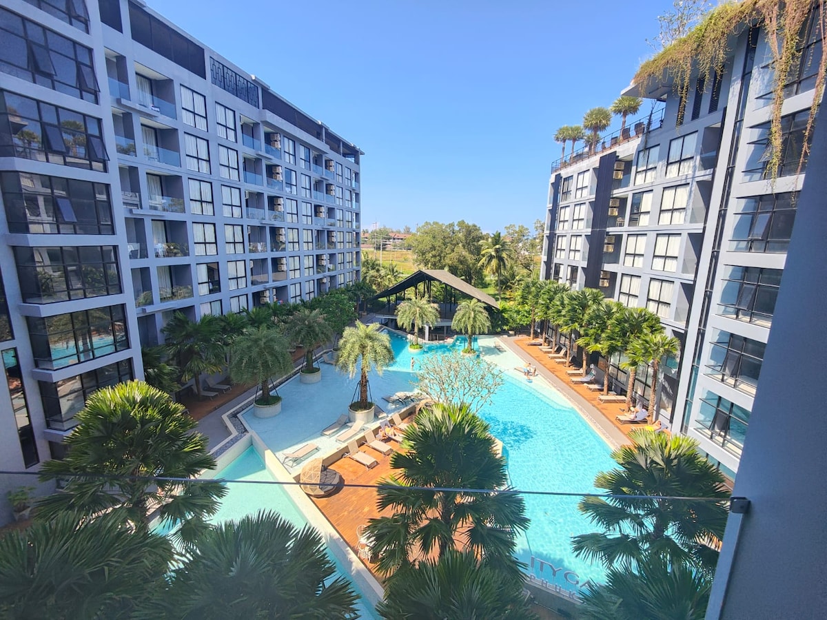 A spacious courtyard showcases three swimming pools surrounded by lush tropical landscaping. Modern buildings frame the area, reflecting sunlight through large windows. Palm trees and lounge areas provide inviting spaces for relaxation under clear blue skies.