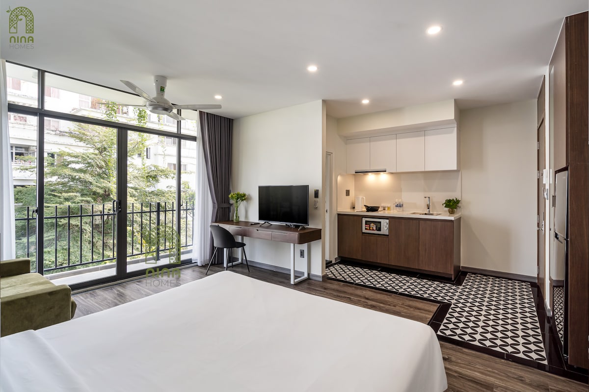 A well-lit studio features a spacious bed in the foreground with crisp white linens. A kitchenette is visible, complete with modern appliances and a dining area. Large glass doors open to a private balcony, offering views of greenery. A comfortable seating area is positioned nearby.
