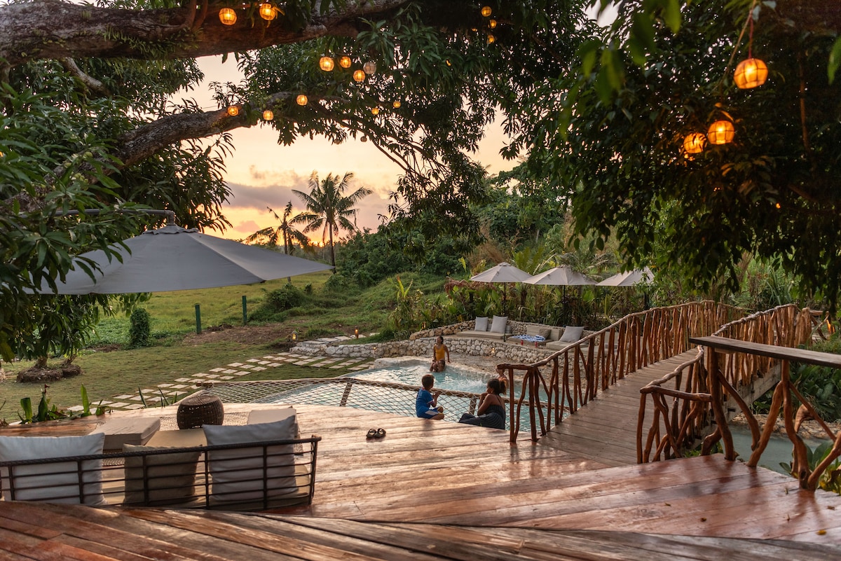 An outdoor area is seen with wooden decking leading to a swimming pool, surrounded by lush greenery. Lounge chairs with umbrellas and decorative lights hang from above, casting a warm glow. A serene atmosphere is established by the sunset in the background.