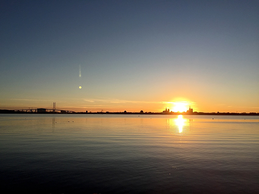 The image captures a calm body of water reflecting a vibrant sunset. The sun, partially below the horizon, casts warm hues across the sky and water. Silhouettes of distant buildings and structures can be seen along the shore, enhancing the serene landscape.