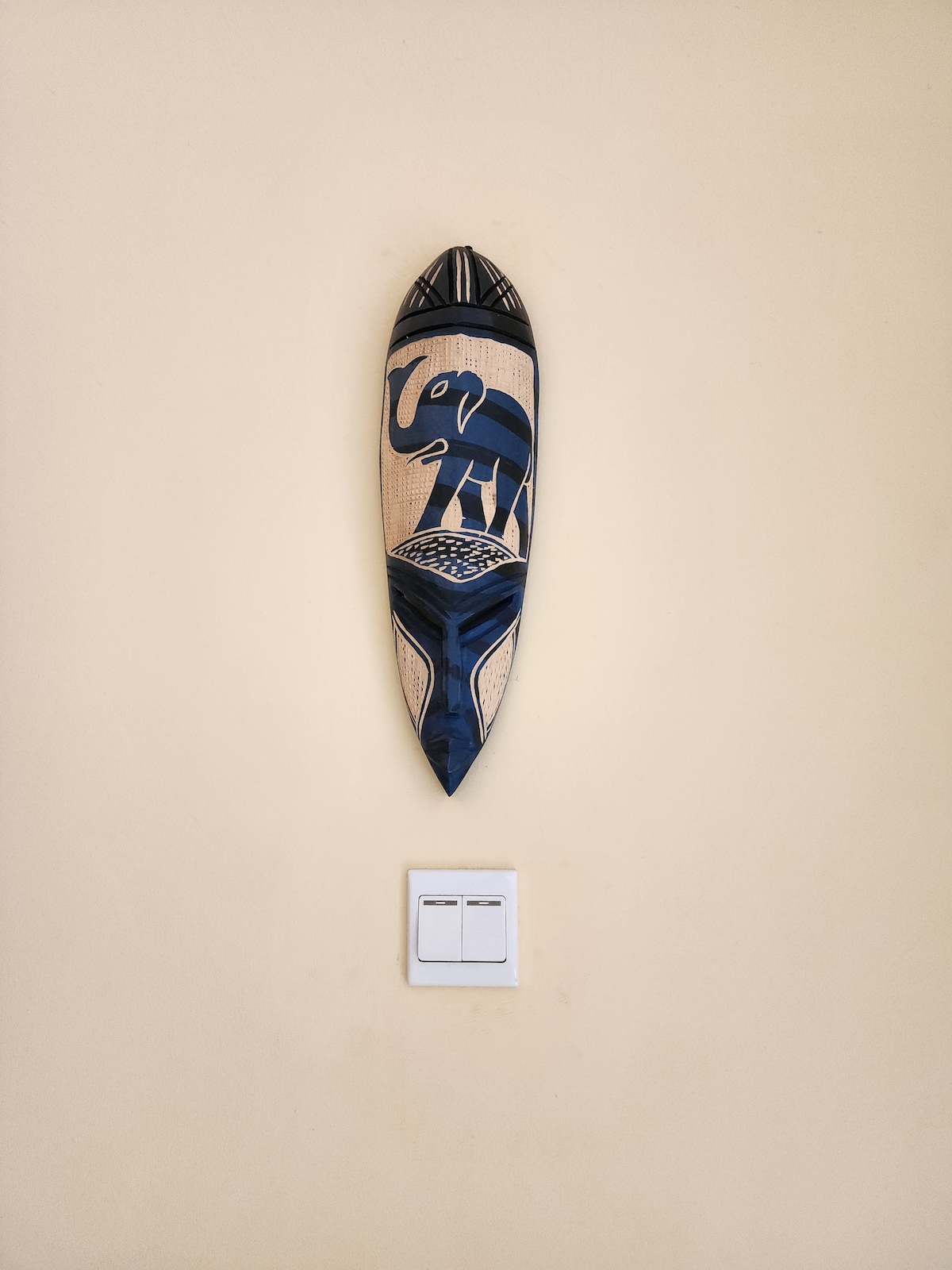 A decorative wall mask featuring an elephant design is mounted against a beige background. Below, a light switch panel with two buttons is visible, accentuating the simplicity of the space.