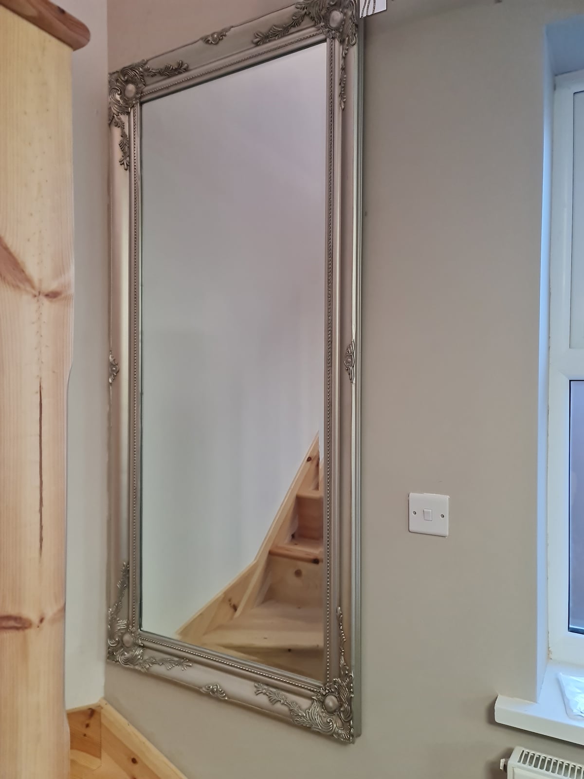 A tall, ornate mirror is mounted on the wall, reflecting the light and enhancing the spaciousness of the area. In the background, a glimpse of wooden stairs can be seen leading to the upper level, with natural light coming in through a nearby window.