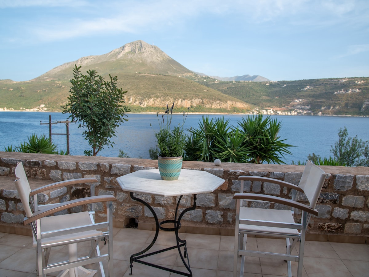 A small outdoor seating area features a round marble table surrounded by two white chairs. Lush greenery and a potted plant are visible on the table, with a scenic view of a mountainous landscape and calm waters in the background.