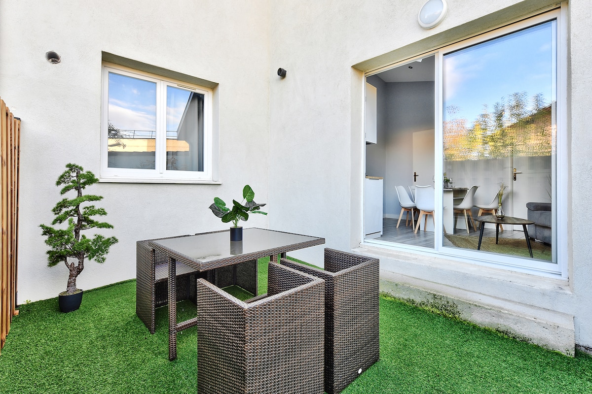 A private terrace features a green artificial lawn with a table and four chairs. A small decorative plant is placed on the table, while a slender tree stands in a pot nearby. Large glass doors lead into the living area.