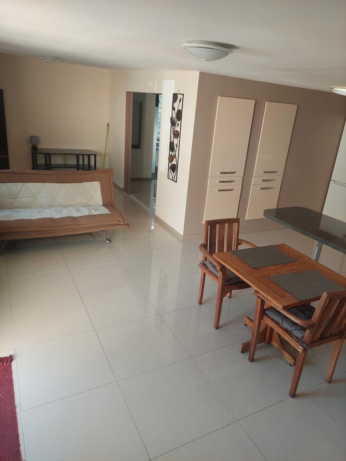 An open living space features a light-colored tiled floor with a small dining area set for four. A brown sofa bed occupies one side, while a kitchenette is positioned nearby. Natural light fills the room through adjacent doorways, contributing to a bright atmosphere.
