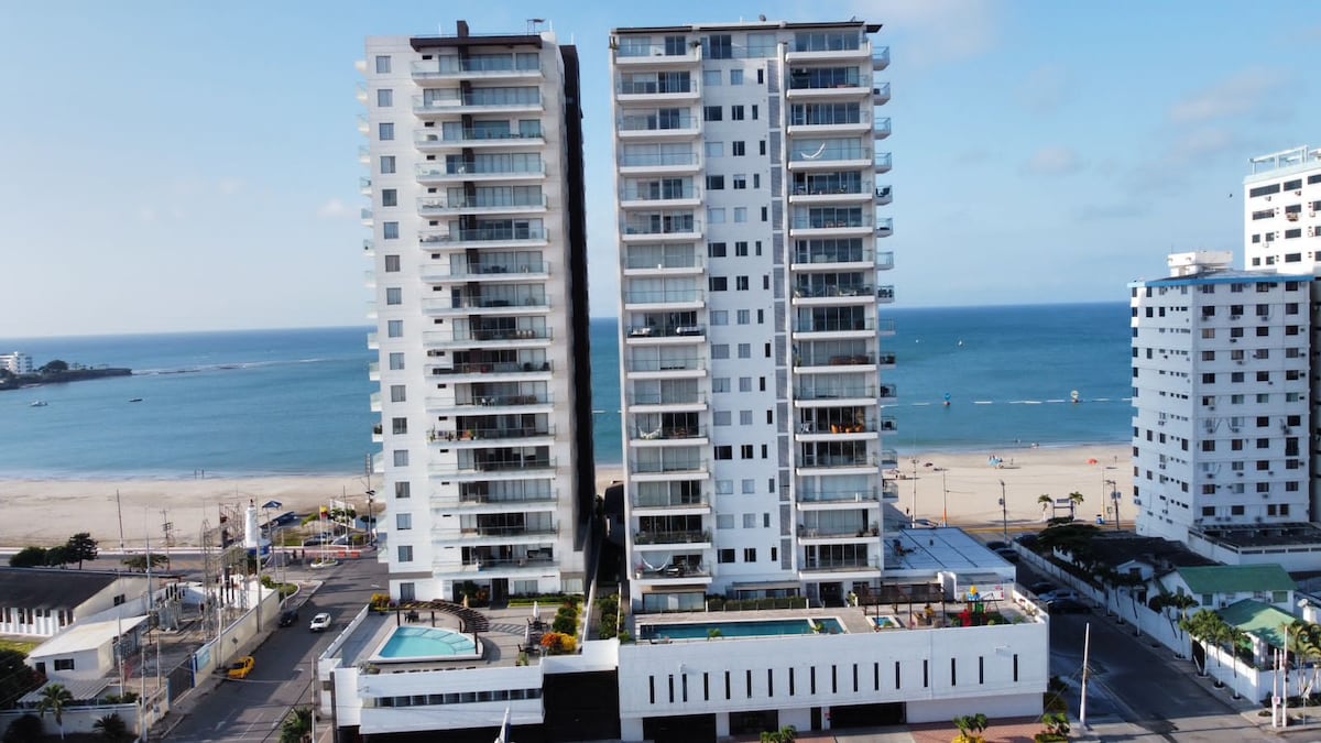 Two modern buildings stand adjacent to a sandy beach, offering scenic ocean views. Each structure features balconies, with some units overlooking a shared pool area. The beach is visible in the distance, highlighting a tranquil coastal setting.