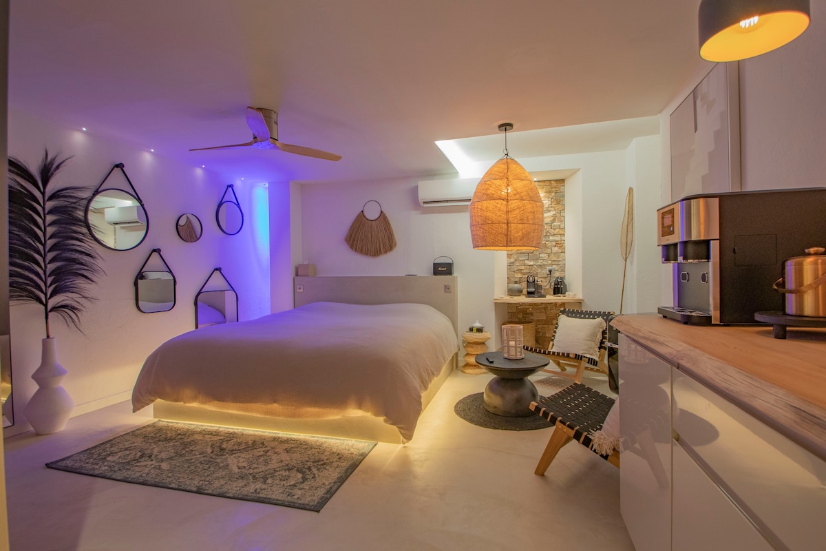 A spacious room features a large bed with soft bedding, complemented by unique mirrors and decorative elements. Ambient lighting adds a soothing touch, while a stone wall provides texture. A seating area with modern furnishings is also visible, creating a relaxing atmosphere.