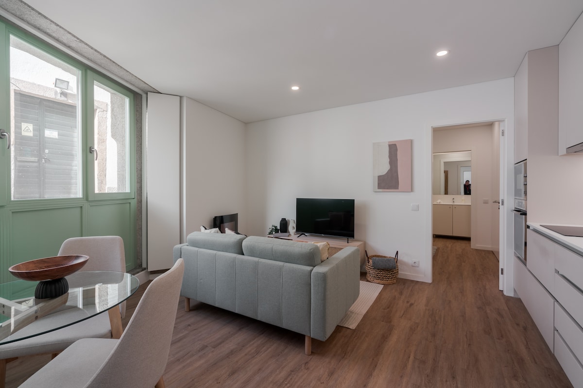 A modern open living area features a light grey sofa facing a flat-screen television. A dining table with white chairs is situated nearby. Natural light enters through large windows, highlighting the warm wood flooring. A doorway leads to the bathroom and bedroom, visible in the background.