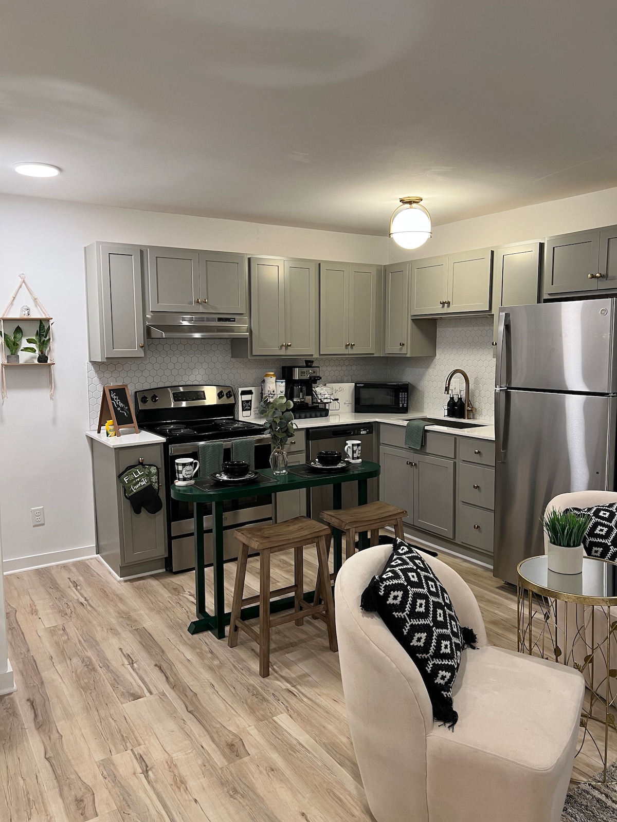 A modern kitchen area features gray cabinetry and sleek appliances, including a stainless-steel refrigerator and oven. A small dining table with two stools is positioned beside a decorative plant, while a neatly set table includes matching dishware. Light streams in from overhead, enhancing the inviting space.