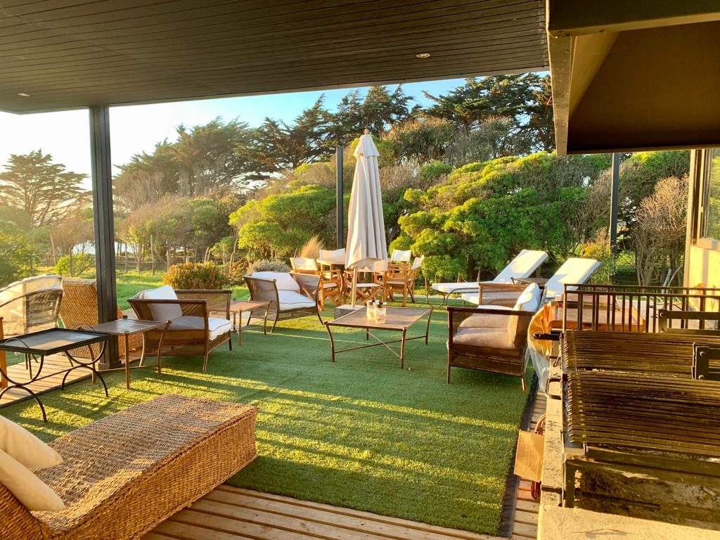 An outdoor terrace is set with comfortable seating arrangements, including wicker chairs and lounge beds. A patio table surrounded by chairs invites gatherings. Lush greenery is visible in the background, while a large umbrella offers shade. A grill is positioned for outdoor cooking.