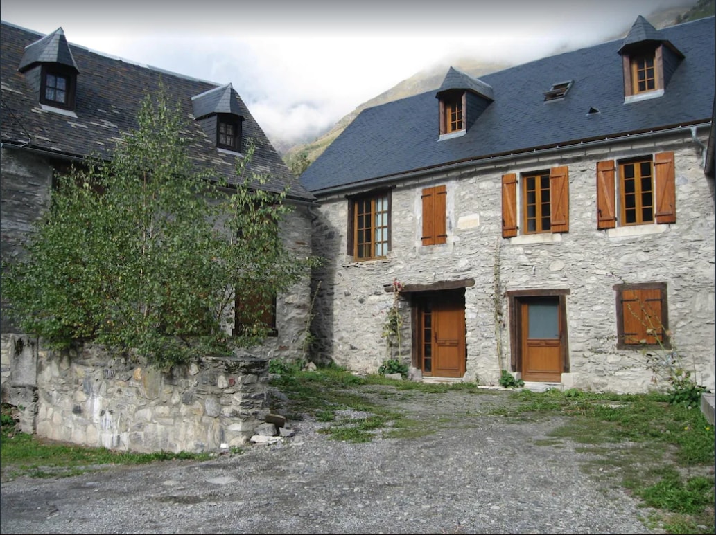 A stone facade features a quaint two-story residence with wooden shutters and a gravel courtyard. Lush greenery is visible near the entrance, and the surrounding mountains can be seen in the background, enhancing the tranquil setting.