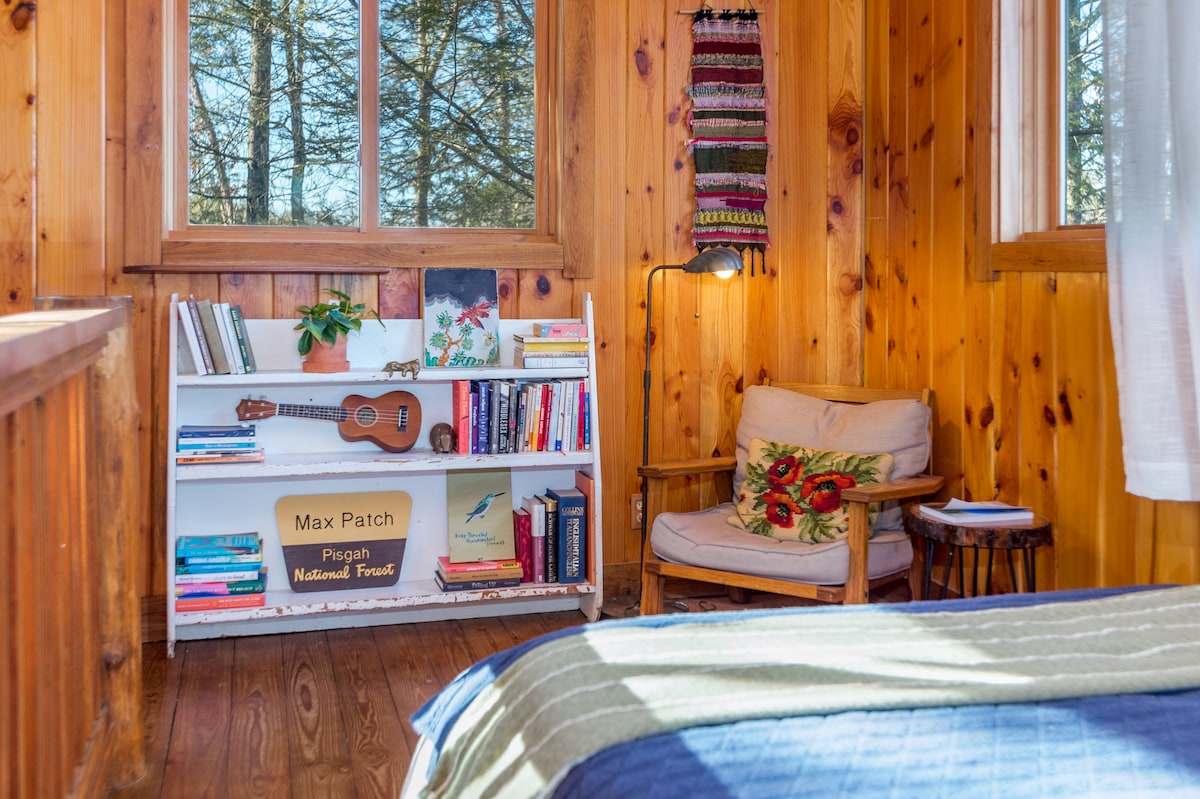 A cozy nook is depicted, showcasing a bookshelf filled with books and a ukulele. A comfortable armchair is placed beside a small side table, inviting relaxation. Natural light filters through a large window, illuminating the warm wooden walls and surrounding greenery.