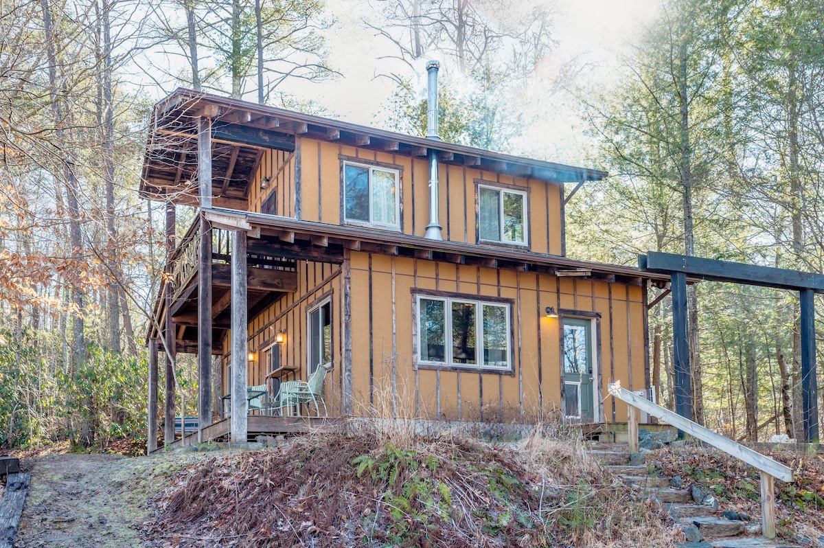 A two-story cabin is set amidst tall trees, featuring a warm wooden exterior and multiple large windows. An inviting front porch with seating provides space for relaxation. Stairs lead up to the main entrance, while a deck extends from the second floor, offering scenic views of the surroundings.