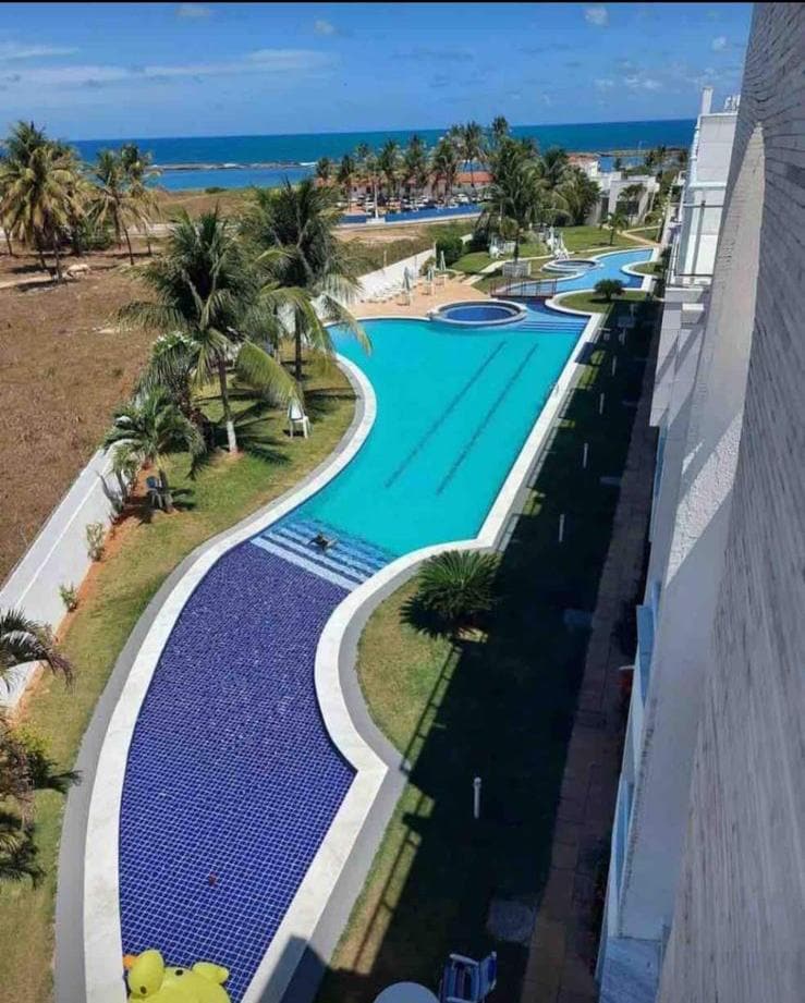 A large outdoor pool with a unique shape is surrounded by lush greenery and palm trees, with ocean views visible in the background. The pool features a light blue hue and is complemented by decorative tile patterns. Sunlight casts gentle shadows across the water.