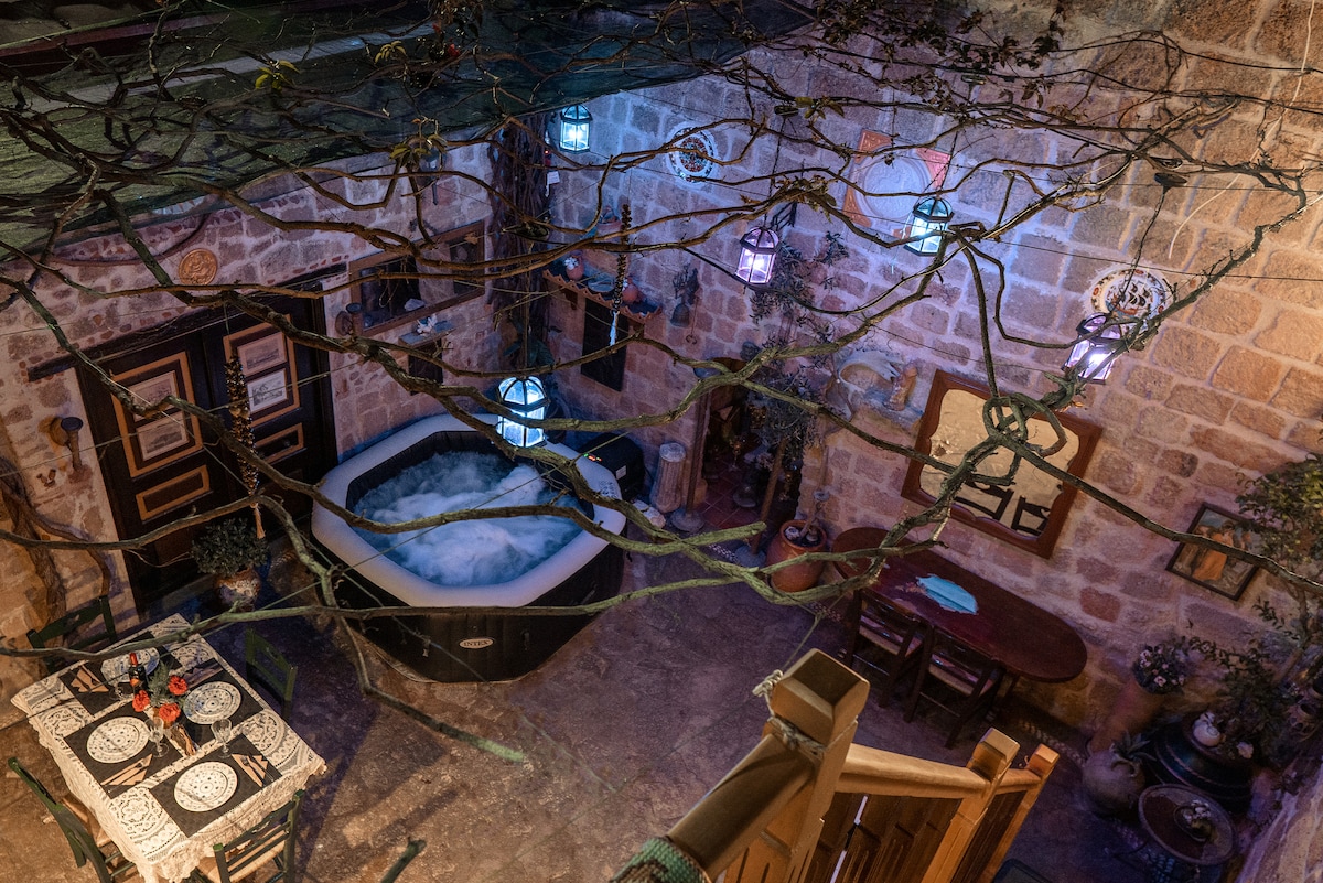 An interior view reveals an outdoor spa with bubbling water, surrounded by stone walls and rustic decor. Lanterns hang from the ceiling, casting a warm glow. A dining table is set nearby, contributing to a cozy and inviting atmosphere.