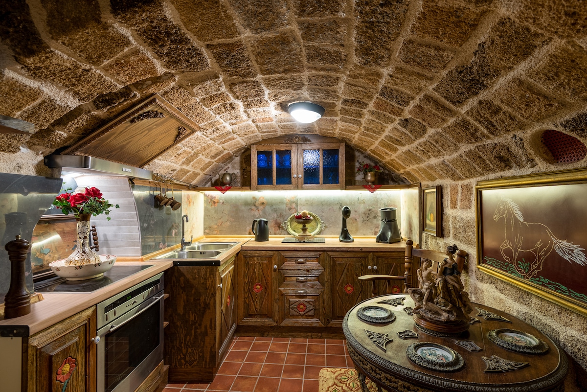 A rustic kitchen showcases vaulted stone ceilings and wooden cabinetry. Understated appliances and decorative elements are arranged harmoniously. A round dining table is placed center stage, adorned with detailed pottery, while soft, ambient lighting enhances the warm tones of the stone walls.