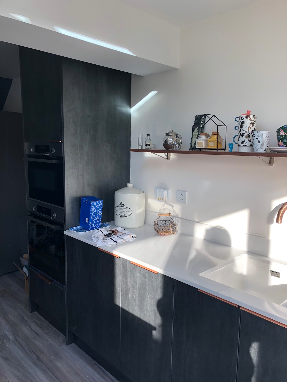 A modern kitchen area features sleek cabinetry and a countertop illuminated by natural light. An array of decorative items is arranged on open shelves, alongside kitchen appliances. A stainless-steel sink is positioned beneath a stylish faucet, creating an inviting workspace for meal preparation.