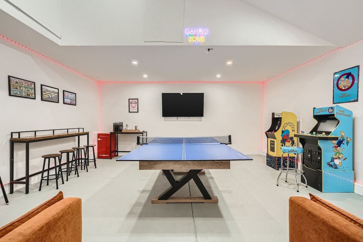 The game room is equipped with a blue ping-pong table at its center. Surrounding elements include a 65” TV mounted on the wall, arcade games, and a bar area with a mini-fridge. LED lights cast a soft glow around the space, enhancing the fun atmosphere.