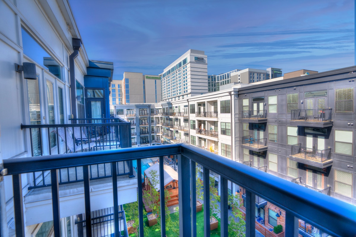 A balcony view showcases the surrounding multifamily complex, featuring modern architecture with a mix of glass and dark siding. An inviting courtyard area with greenery is visible below, while the skyline of nearby buildings stretches in the background under a clear blue sky.