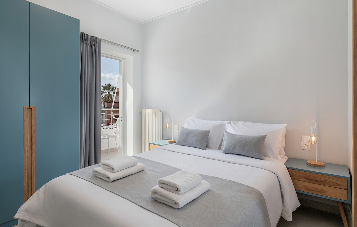 A well-lit bedroom features a spacious bed dressed in crisp linens and accented with soft grey pillows. A wardrobe with blue tones stands against the wall. Natural light filters through the window, which leads to a balcony, enhancing the calming atmosphere.