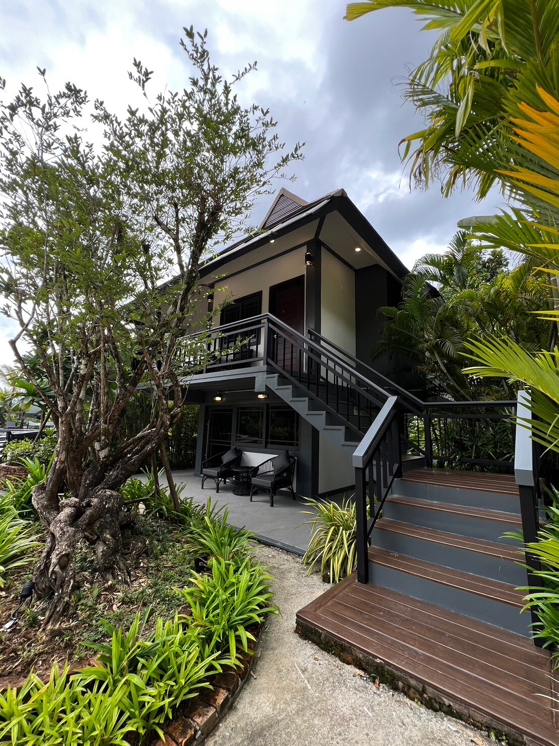 The villa suite's exterior features a two-story structure with a steeply pitched roof. A landscaped path leads to a set of stairs that ascend to a spacious deck with seating. Lush greenery surrounds the home, enhancing the inviting entrance.