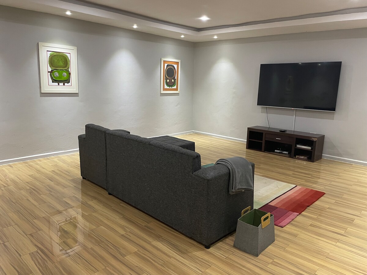 A spacious living area features a dark grey sectional sofa positioned towards a large flat-screen TV mounted on the wall. Two framed artworks are displayed above the sofa, and a soft area rug lies on the wooden floor, adding color to the room.