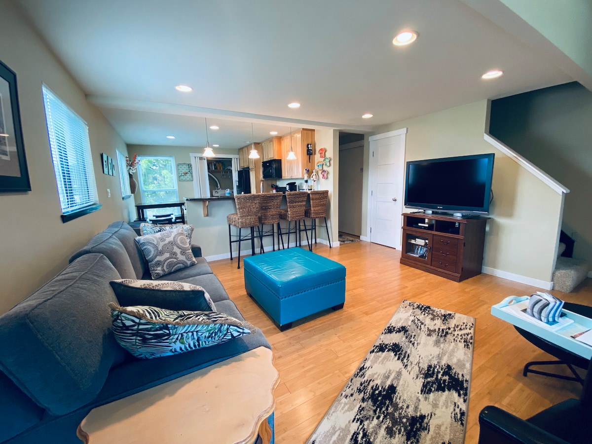 An open living area features a gray sofa adorned with decorative pillows, a teal ottoman, and a patterned area rug. Bar stools surround a kitchen island in the background. A flat-screen TV is positioned on a wooden console, with a staircase visible to the right.