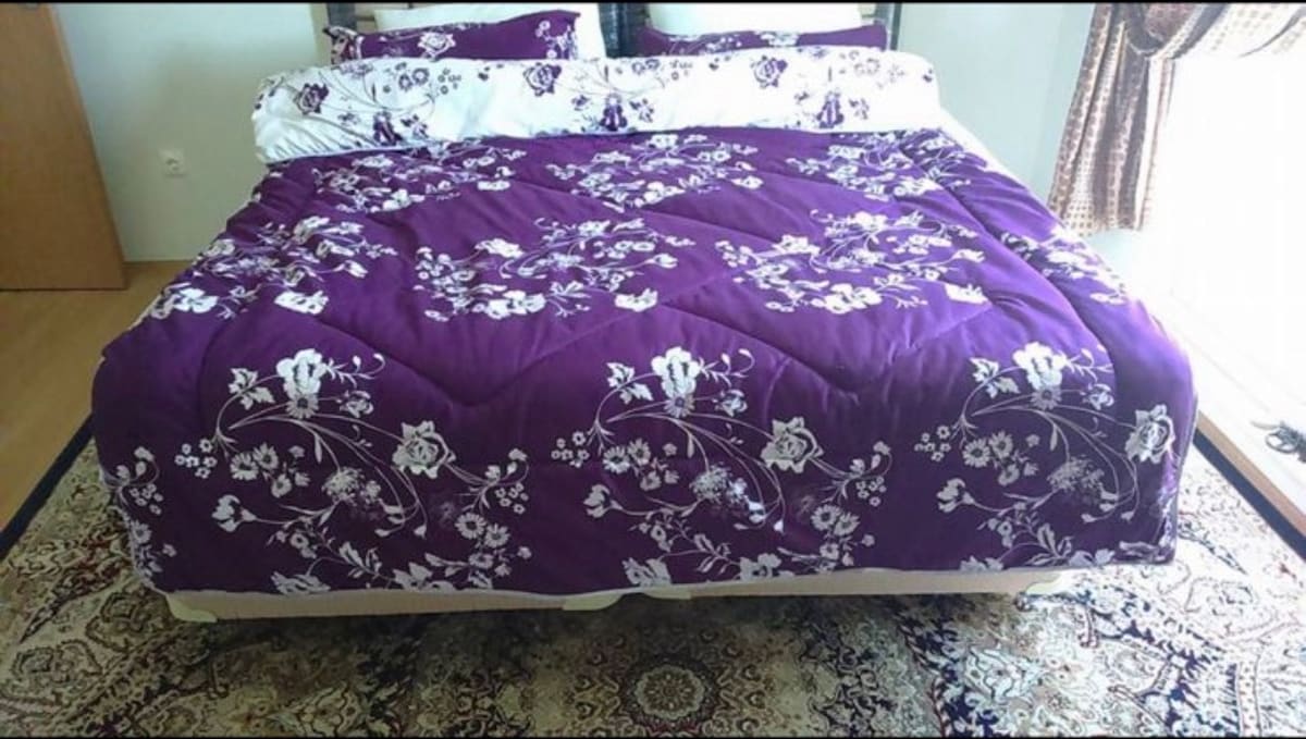 A spacious bed is dressed with a floral purple quilt featuring white accents. Decorative pillows are arranged at the head of the bed. Natural light enters through a nearby window, casting soft shadows across a patterned area rug beneath the bed.
