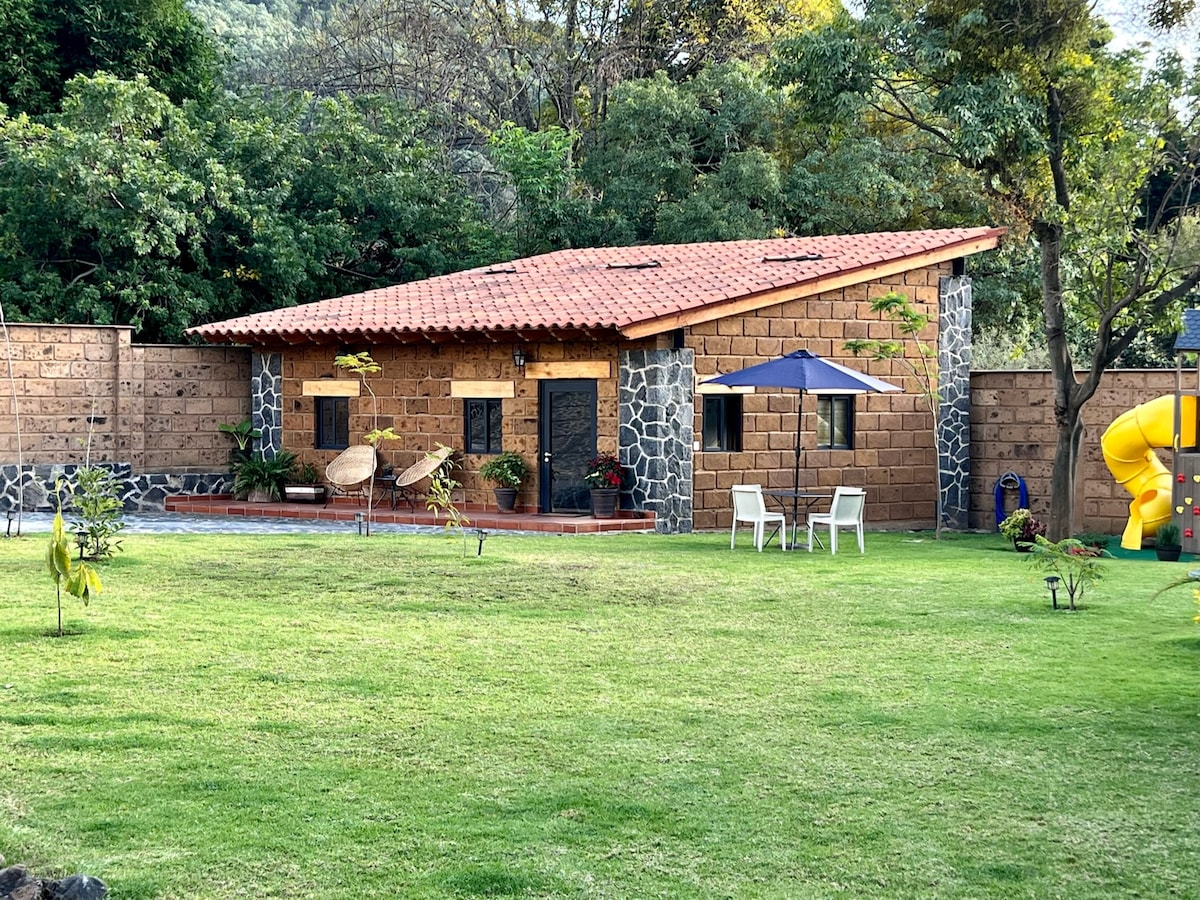 A charming cabin is surrounded by lush greenery, featuring a red-tiled roof and stone accents. The outdoor area includes two white chairs and an umbrella, with a yellow play structure visible to the side. A well-maintained lawn completes the serene setting.