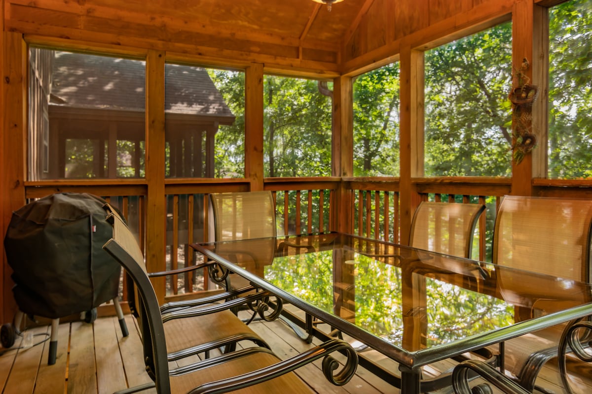 A screened-in back deck features a glass-top dining table surrounded by several chairs. Sunlight filters through the surrounding trees, creating a serene outdoor dining experience. A barbecue grill is present in the corner, enhancing the outdoor living space.