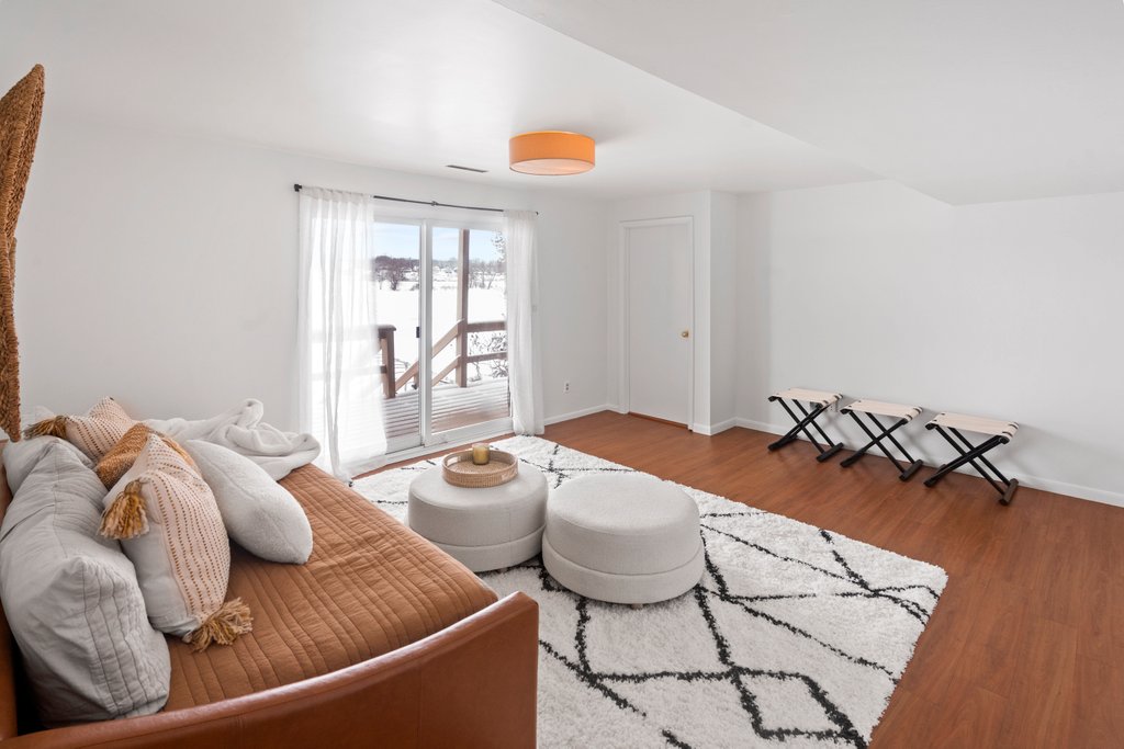 A spacious living area is presented with a cozy daybed adorned with soft pillows. Natural light fills the room through large glass doors, which open to a deck. A round ottoman and a patterned area rug enhance the inviting ambiance.