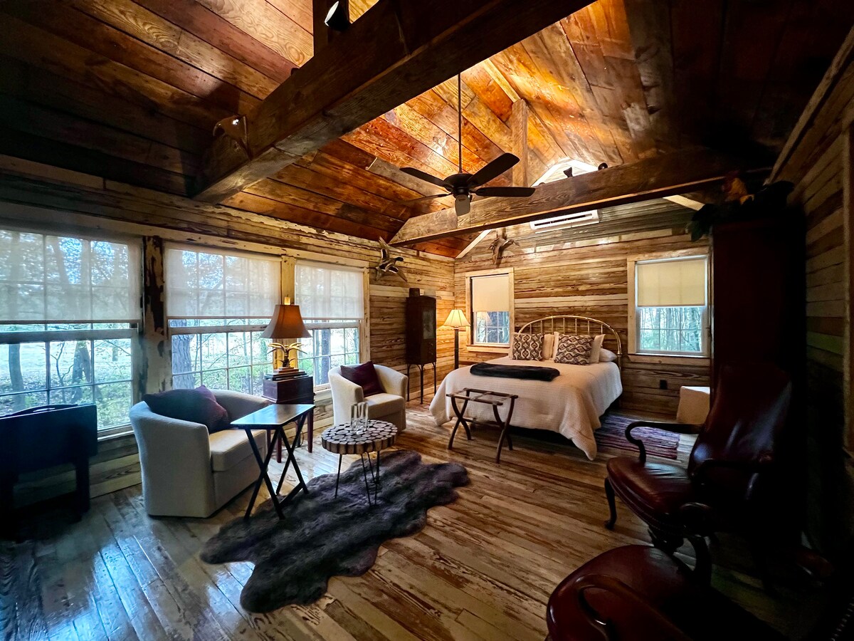 A spacious bedroom features wooden walls and a high ceiling, with natural light streaming through large windows. Two comfortable chairs and a small table are present, along with a cozy bed draped with a dark blanket. A decorative rug adds texture to the wooden floor.
