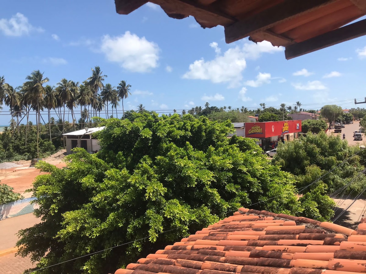 A rooftop view captures the lush greenery of palm trees against a clear blue sky. A vibrant street scene is visible with nearby commercial buildings, showcasing a blend of nature and urban life. The terracotta roofing adds warmth to the perspective.