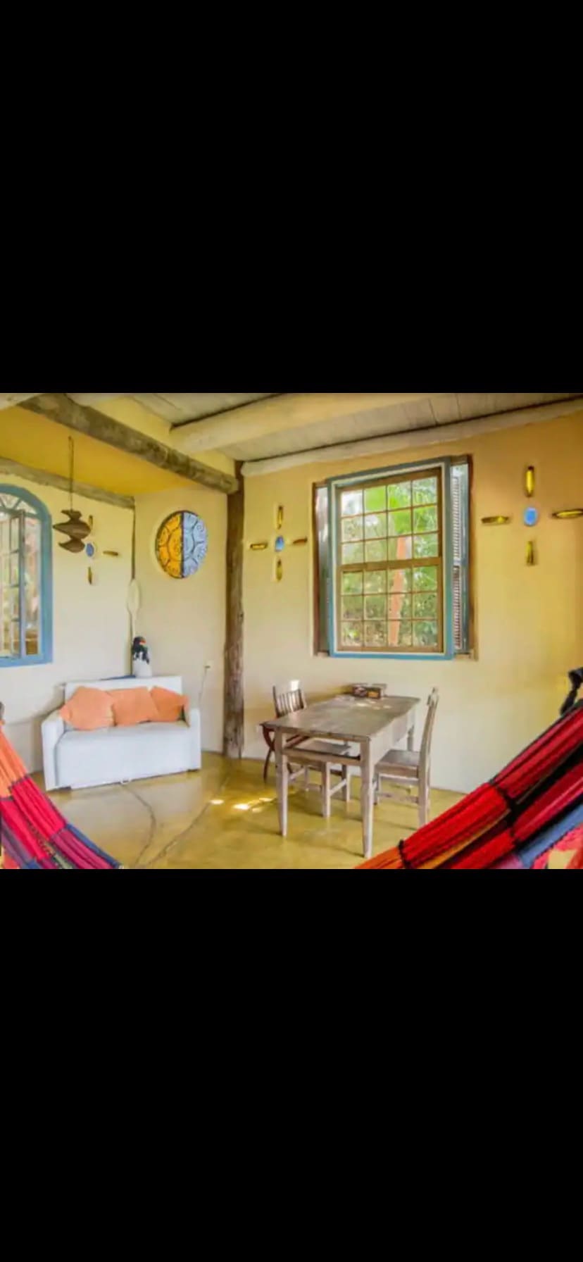 A cozy sitting area features a white sofa adorned with orange cushions and a small wooden table. Decorative elements include colorful wall art and a round window, allowing natural light to illuminate the space. A pair of vibrant hammocks hangs nearby.