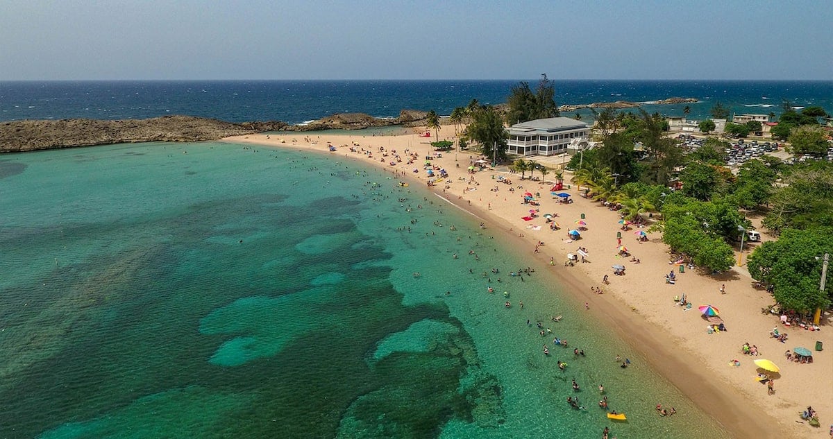 A coastal view presents a blend of golden sand and turquoise waters, dotted with beachgoers enjoying the sun. Lush greenery lines the shore, and a modest building is visible in the background. Gentle waves lap against the beach, creating a serene atmosphere.