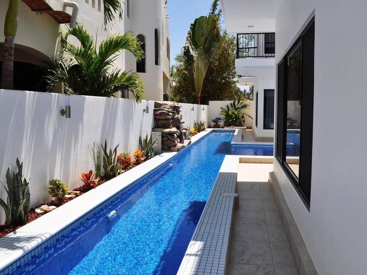 A serene outdoor pool area features a long, narrow pool lined with vibrant greenery and well-maintained landscaping. Sunlight reflects off the clear blue water, while the adjacent white walls provide a clean contrast. The space is bordered by tropical plants, creating a tranquil atmosphere.