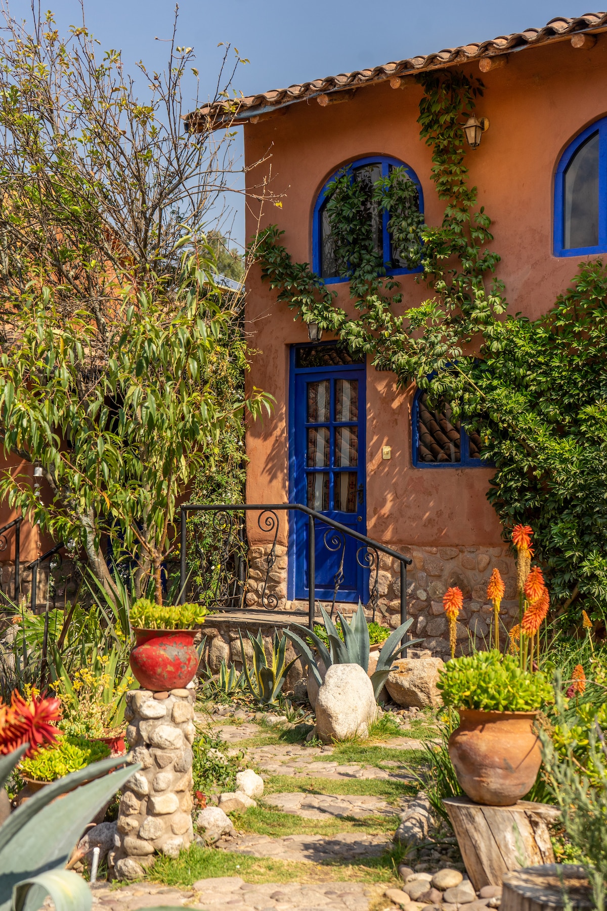 A charming entrance is framed by vibrant foliage and colorful flowers, leading to a warm-hued building with distinctive blue windows. The stone pathway guides visitors toward the door, inviting exploration of the lush surroundings and well-maintained garden.
