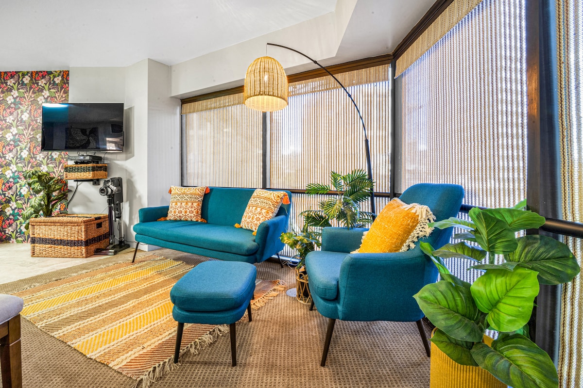 A vibrant living area is filled with plush seating and decorative accents. A teal couch and matching armchair offer comfort, with a cozy ottoman inviting relaxation. Large windows allow natural light to filter through bamboo shades, highlighting decorative plants and a warm woven rug underfoot.