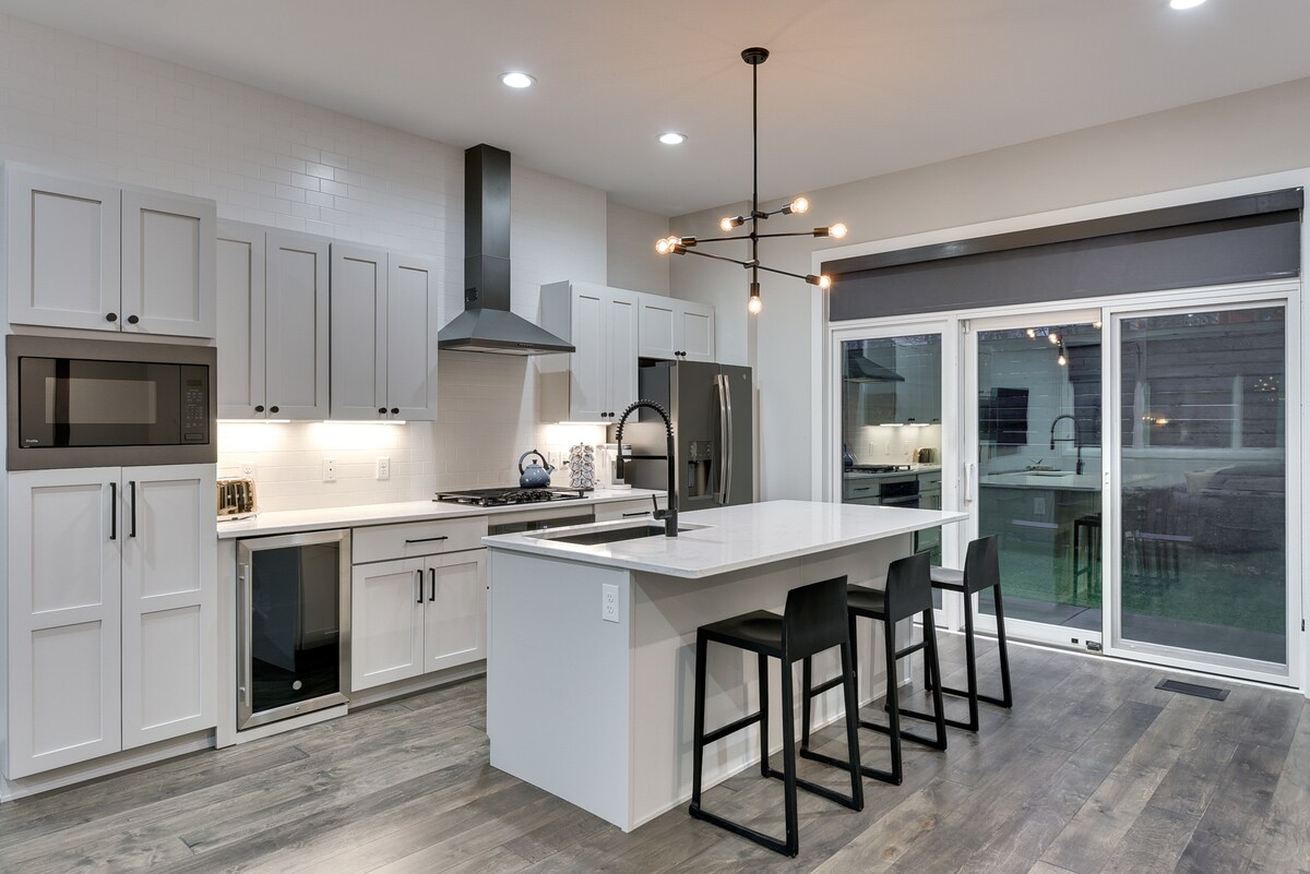 An open-concept kitchen is showcased with modern cabinetry and stainless steel appliances. A central island with seating accommodates six black bar stools. Large sliding glass doors provide access to an outdoor space, and stylish pendant lighting hangs above the island.