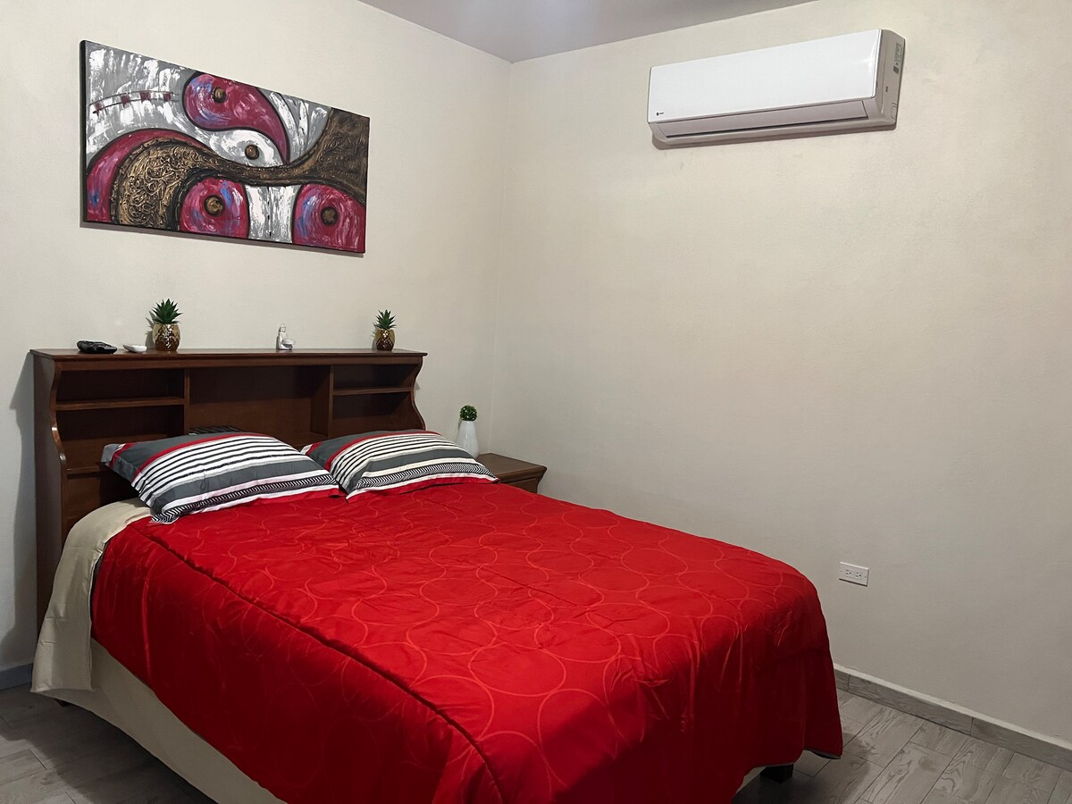 A well-lit sleeping area features a spacious bed with a bright red coverlet and striped accent pillows. A wooden headboard provides support to the bed, while a wall-mounted air conditioning unit is visible above. A piece of artwork adds color to the wall.