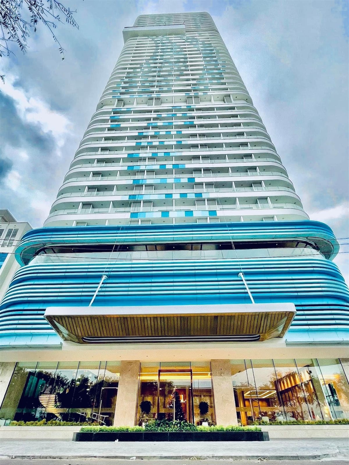A tall modern building with an eye-catching, wavy turquoise façade is shown. The ground level features large glass doors, inviting entry, and an open lobby area surrounded by greenery. The sky above displays a mix of clouds, enhancing the building's contemporary design.