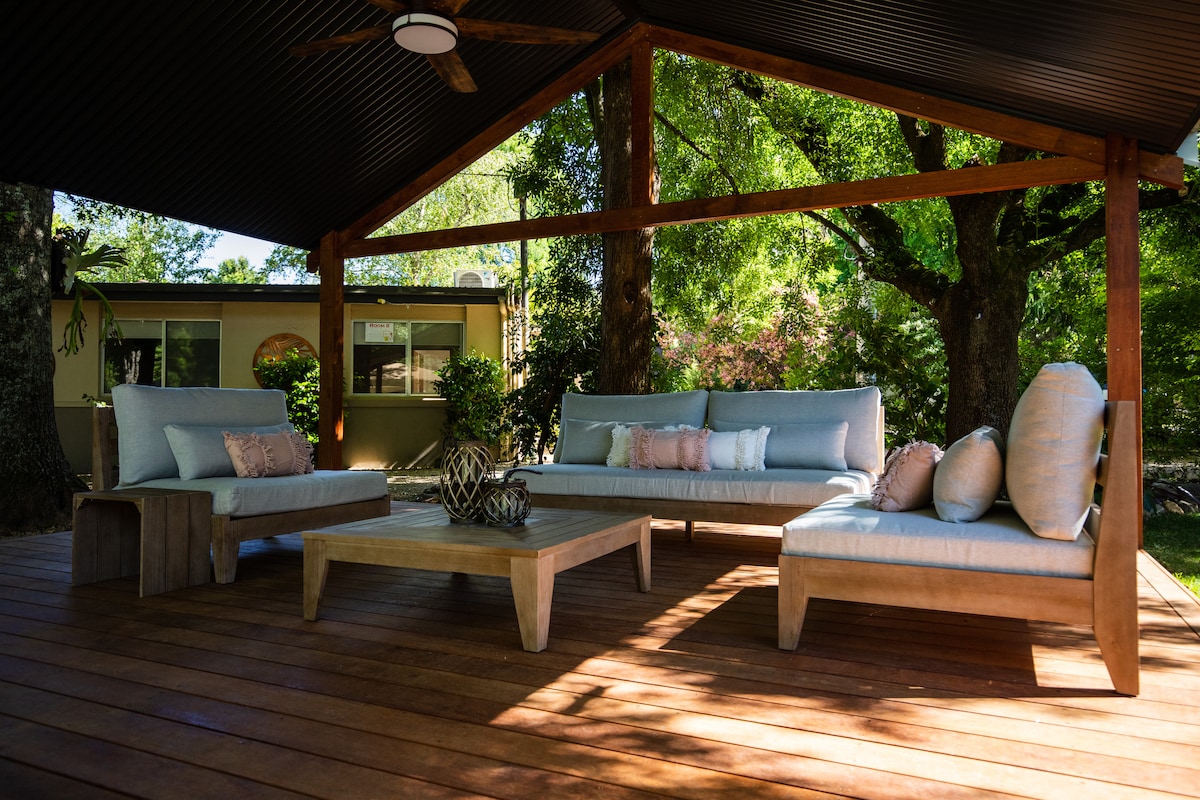 A spacious outdoor seating area features modern lounges and a coffee table, all set on a wooden deck. Lush greenery surrounds the space, complemented by a ceiling fan above and natural light filtering through the trees.