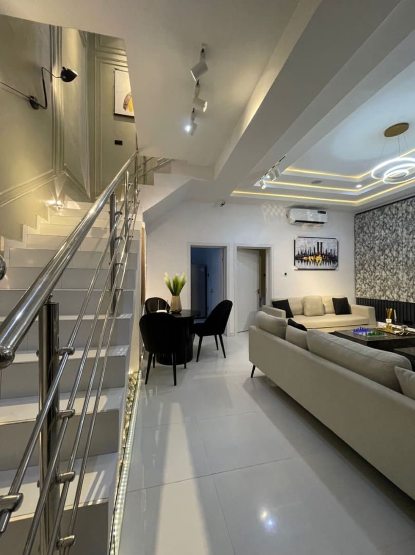 A modern living space features a sleek staircase leading to an upper level. A contemporary seating area with a neutral sofa and dark dining chairs is visible. Soft lighting highlights the elegant decor, while a small table displays fresh flowers in the corner.