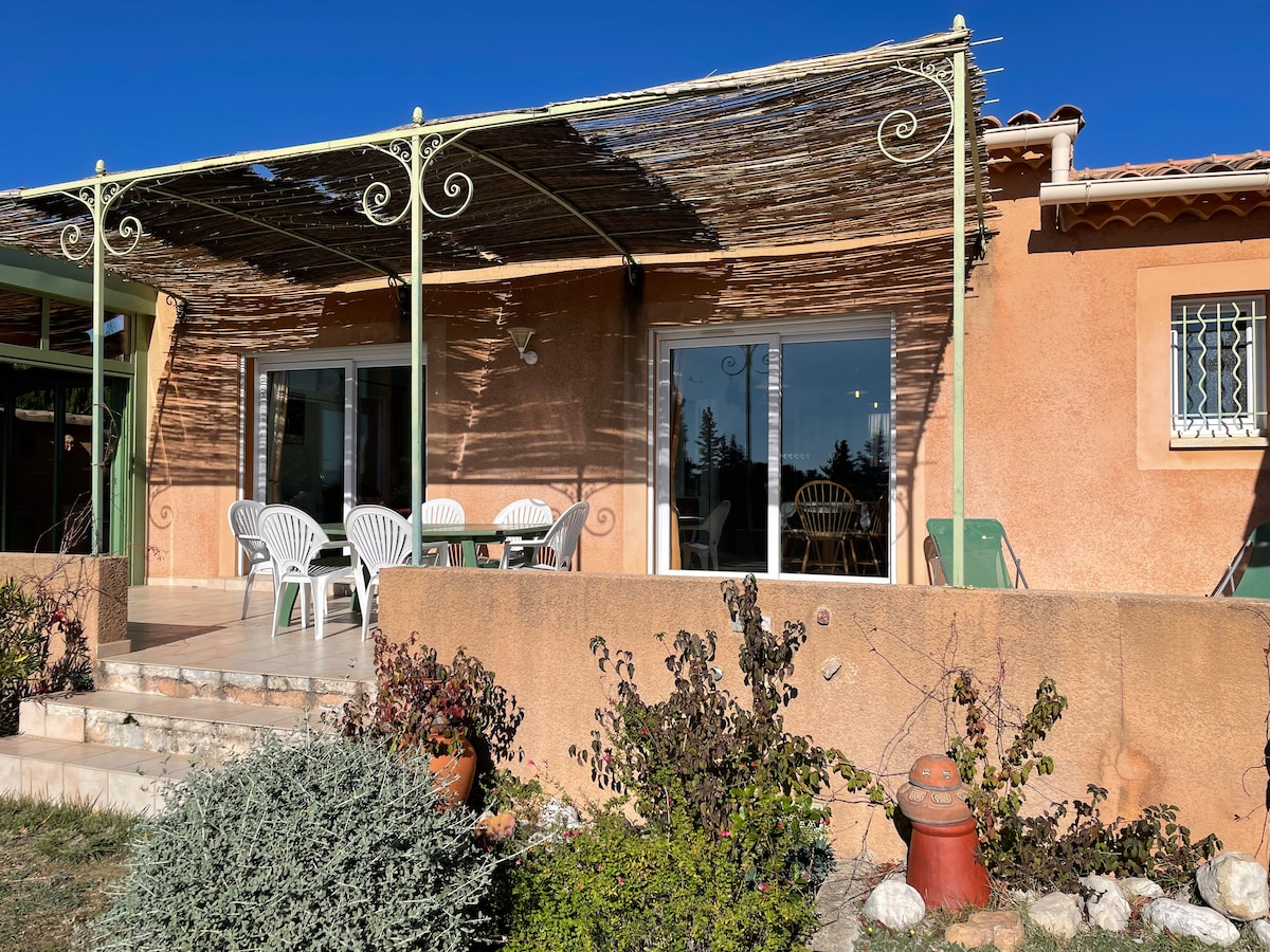 A spacious terrace features a covered seating area with several white chairs, inviting relaxation. The exterior walls are finished in warm tones, complemented by lush greenery and decorative plants. Large glass doors provide a view of the interior, allowing natural light to enhance the space.