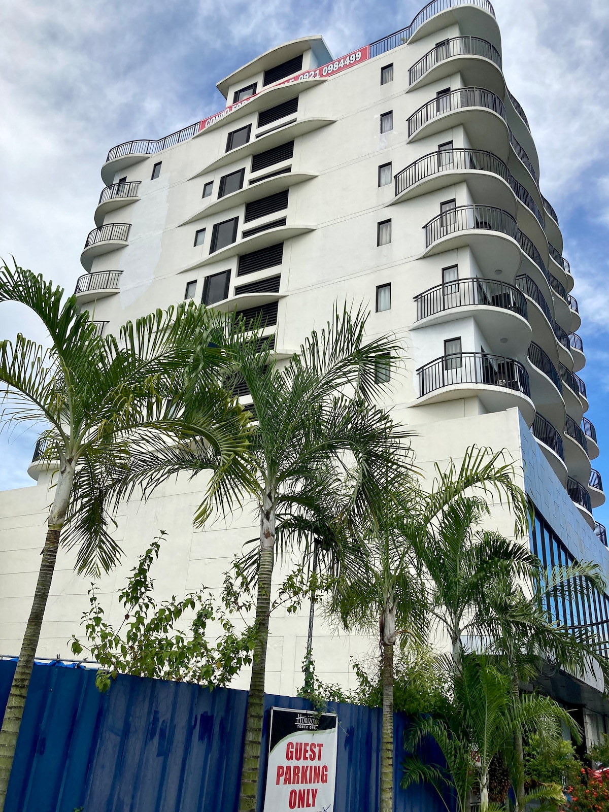 A modern multi-story building is depicted, with a white exterior and multiple balconies. Lush palm trees are visible in the foreground, enhancing the setting. A sign indicating 'Guest Parking Only' is placed at the base of the structure, contributing to the welcoming atmosphere.