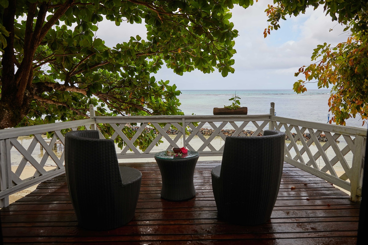 A shaded outdoor seating area features two woven chairs arranged around a small round table, set against a backdrop of the sea. Lush greenery frames the view, and the calm water is visible in the distance, enhancing the serene setting.
