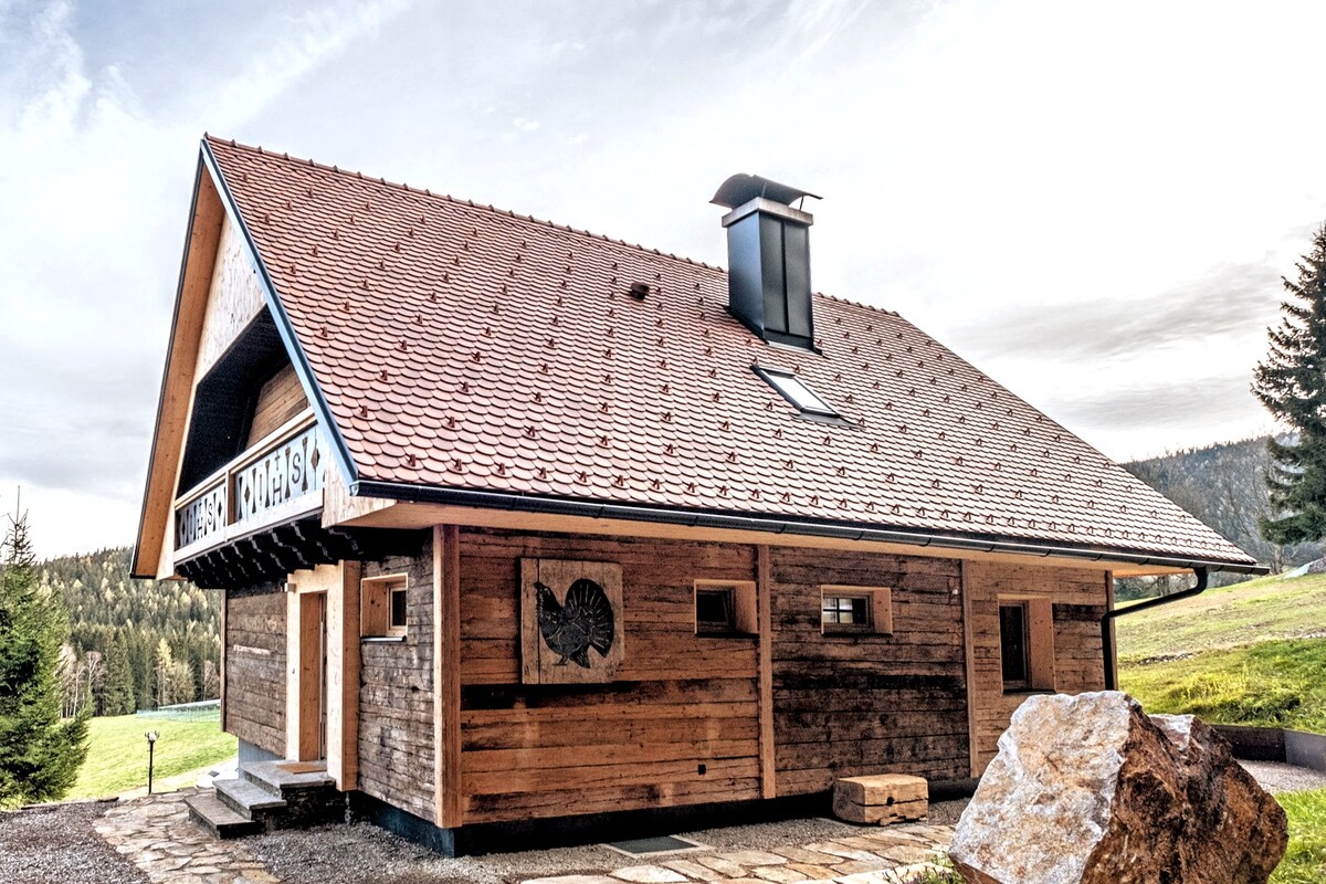 The exterior of the wooden chalet features a sloped roof with traditional red tiles and an elegantly designed chimney. Natural wood tones are highlighted throughout, complementing the surrounding greenery and stone pathway leading to the entrance.