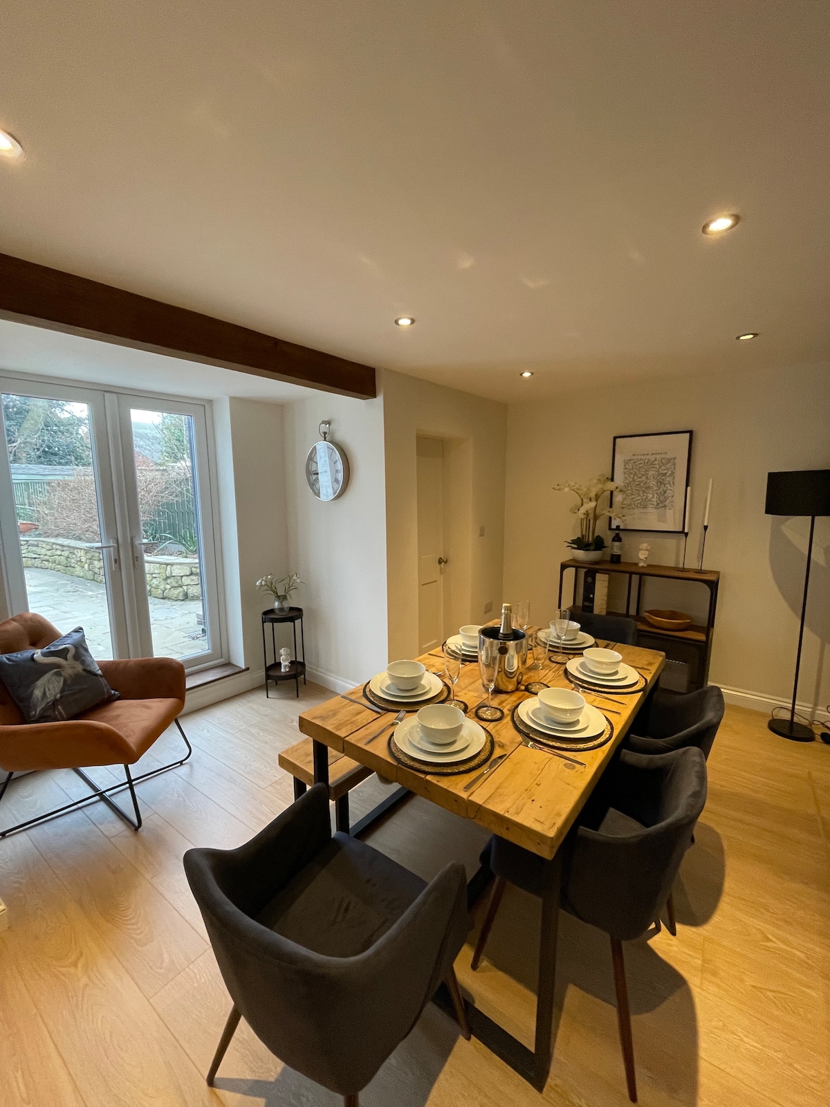 A dining area features a wooden table set with dishes and glassware. Comfortable chairs surround the table, and a cozy armchair is placed nearby. Natural light fills the space through large glass doors, which open to a view of the outdoor area.