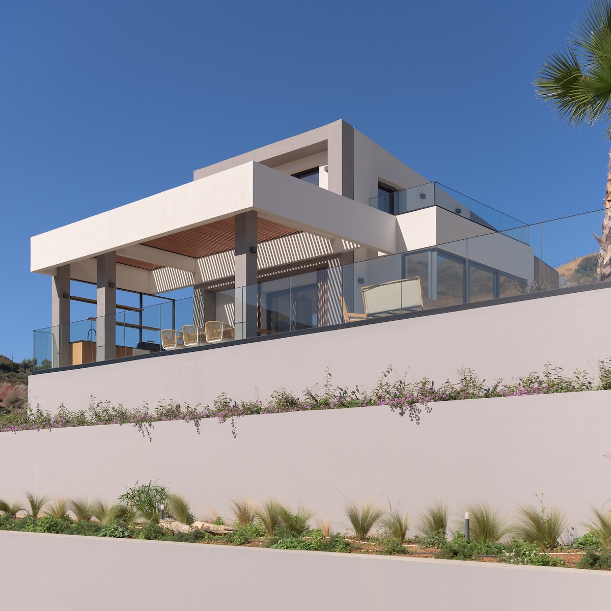 The modern villa is showcased from an elevated angle, featuring clean lines and a minimalist design. A spacious balcony with glass railings offers a view of the surrounding landscape. Lush greenery and ornamental plants adorn the terrace, enhancing the villa's elegant appearance.