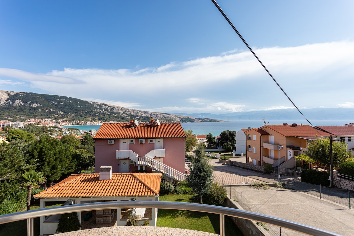 A scenic view from the balcony reveals a coastal landscape with a blend of colorful houses and lush greenery. The azure sea stretches into the horizon, while the distant hills frame the picturesque setting.
